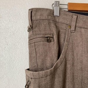 May include: Close-up of brown herringbone-patterned pants with a zipper pocket and a standard pocket. The pants are hanging on a silver hanger. The fabric has a textured appearance.