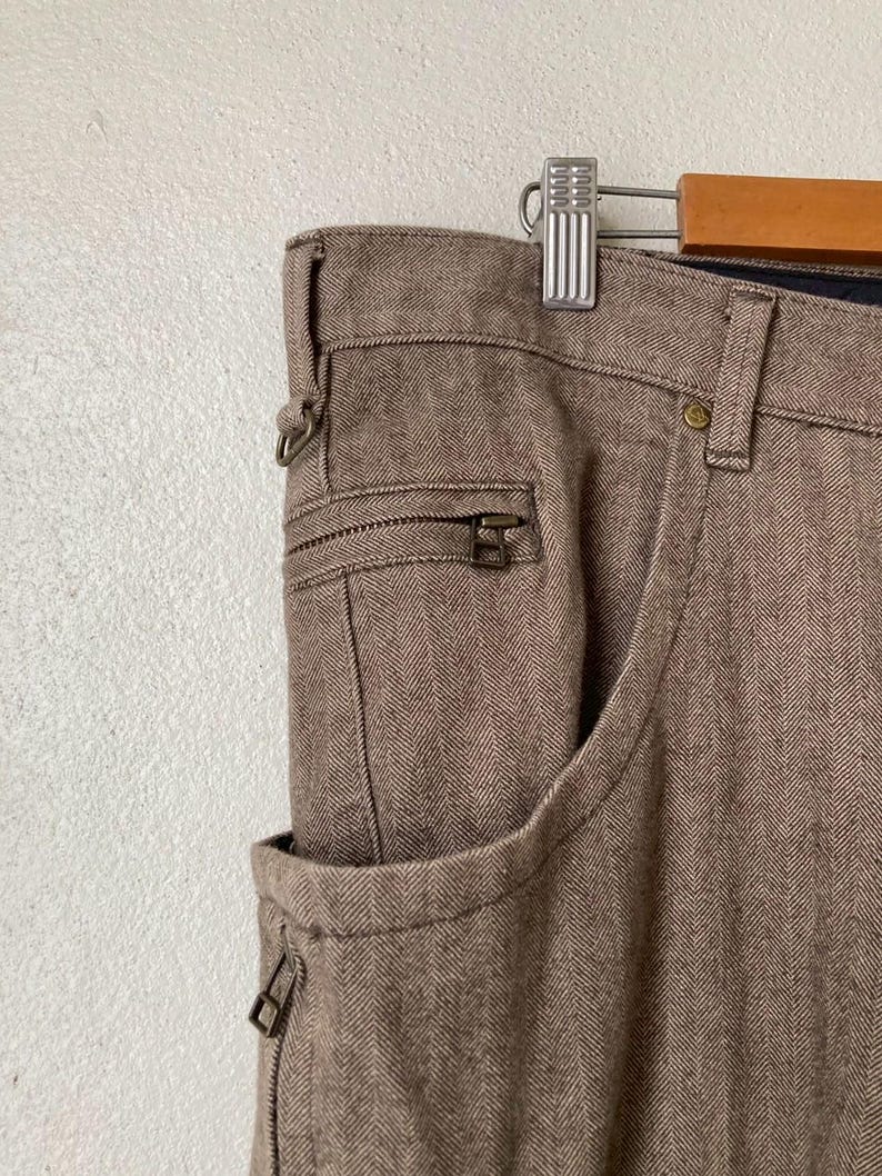 May include: Close-up of brown herringbone-patterned pants with a zipper pocket and a standard pocket. The pants are hanging on a silver hanger. The fabric has a textured appearance.