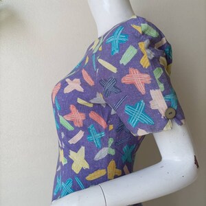 May include: A purple dress with a colorful abstract print. The dress has short sleeves and a button on the cuff.