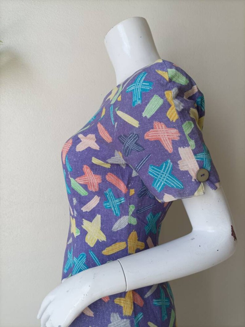 May include: A purple dress with a colorful abstract print. The dress has short sleeves and a button on the cuff.