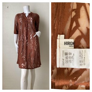 May include: A knee-length, rust-coloured dress featuring a semi-sheer, burnout-style leaf pattern, a V-neckline, and elbow-length sleeves. The interior tag displays the brand name HIROKO and includes care instructions with symbols and Japanese text.