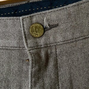 May include: Close-up of gray herringbone trousers with a button that reads "Edwin Golf". The trousers have a dark blue waistband and a button closure. The fabric has a textured appearance.