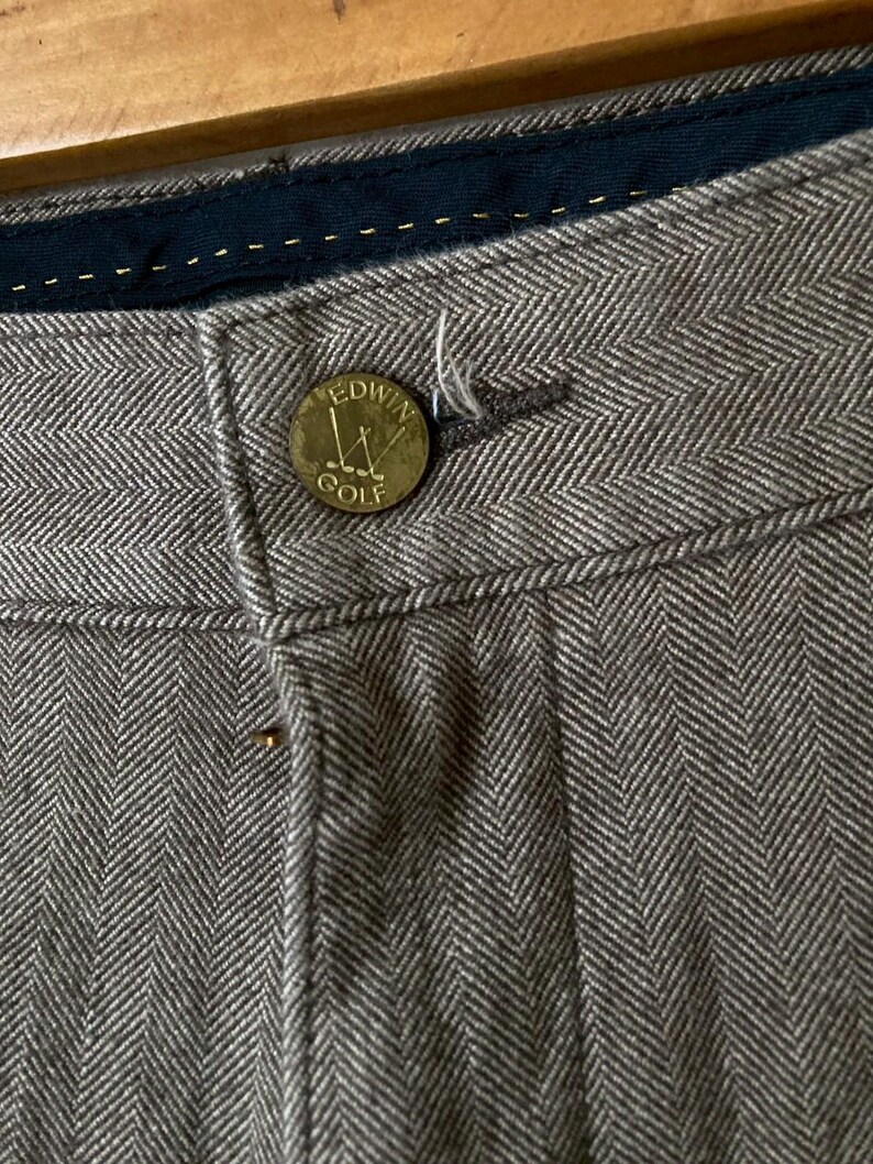 May include: Close-up of gray herringbone trousers with a button that reads "Edwin Golf". The trousers have a dark blue waistband and a button closure. The fabric has a textured appearance.