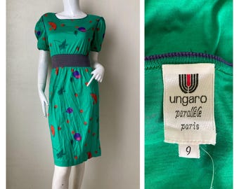 Puede incluir: Un vestido verde vintage con cuello redondo y mangas cortas abullonadas. El vestido presenta una cinturilla gris oscuro y un estampado de formas rojas, moradas y verdes. La etiqueta dice "ungaro parallèle paris".