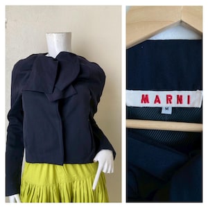 Pode incluir: Um casaco azul marinho com um design único em forma de laço no decote. O casaco é curto e tem mangas compridas. A etiqueta na imagem diz "MARNI" em letras vermelhas. O casaco é combinado com uma saia verde limão.