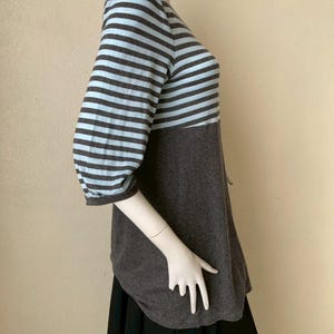 Puede incluir: Una blusa a rayas grises y azul claro con mangas tres cuartos. La blusa tiene una secci&oacute;n inferior gris lisa y un dobladillo redondeado. Las mangas son de rayas horizontales azul claro y grises.