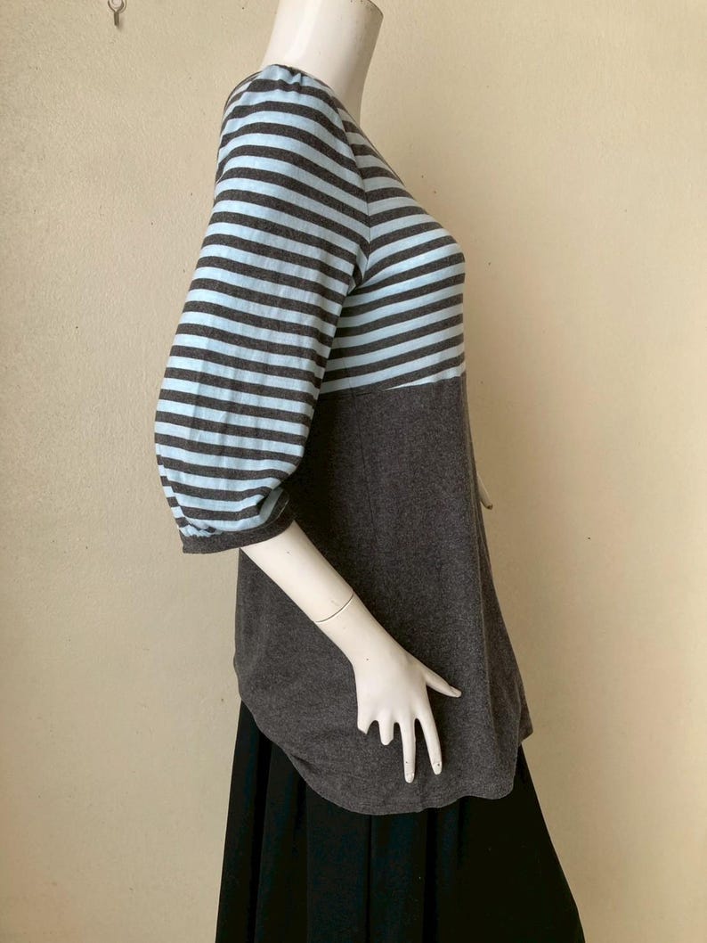 Puede incluir: Una blusa a rayas grises y azul claro con mangas tres cuartos. La blusa tiene una secci&oacute;n inferior gris lisa y un dobladillo redondeado. Las mangas son de rayas horizontales azul claro y grises.