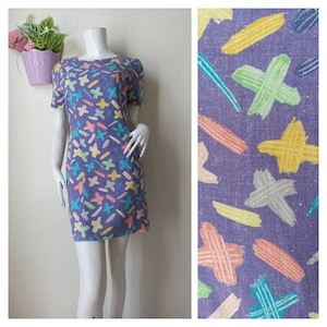 May include: A purple mini dress with a colorful abstract print of stars, lines, and crosses. The dress has short sleeves and a round neckline.