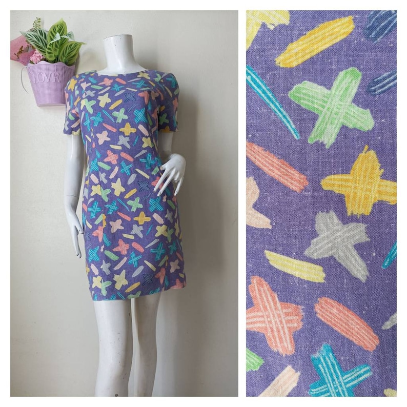 May include: A purple mini dress with a colorful abstract print of stars, lines, and crosses. The dress has short sleeves and a round neckline.