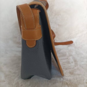 May include: A gray leather satchel bag with a brown leather strap and trim. The bag has a flap closure and a small pocket on the front.