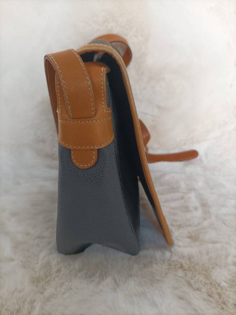 May include: A gray leather satchel bag with a brown leather strap and trim. The bag has a flap closure and a small pocket on the front.