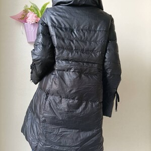 May include: Black, long puffer coat with a high collar. The coat has a quilted design and a flared hem. The sleeves have decorative ties at the cuffs. The coat is displayed on a mannequin.
