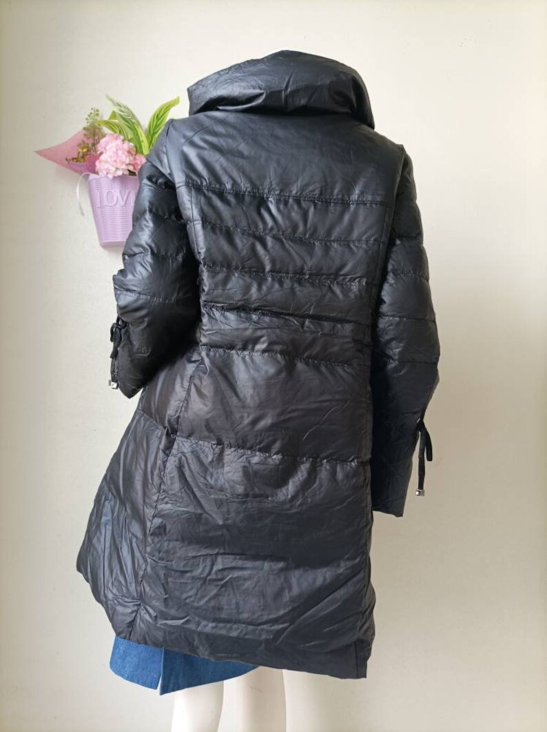 May include: Black, long puffer coat with a high collar. The coat has a quilted design and a flared hem. The sleeves have decorative ties at the cuffs. The coat is displayed on a mannequin.