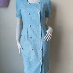May include: A light blue dress with a double breasted button closure and short sleeves. The dress has a pocket on the left side and white buttons.