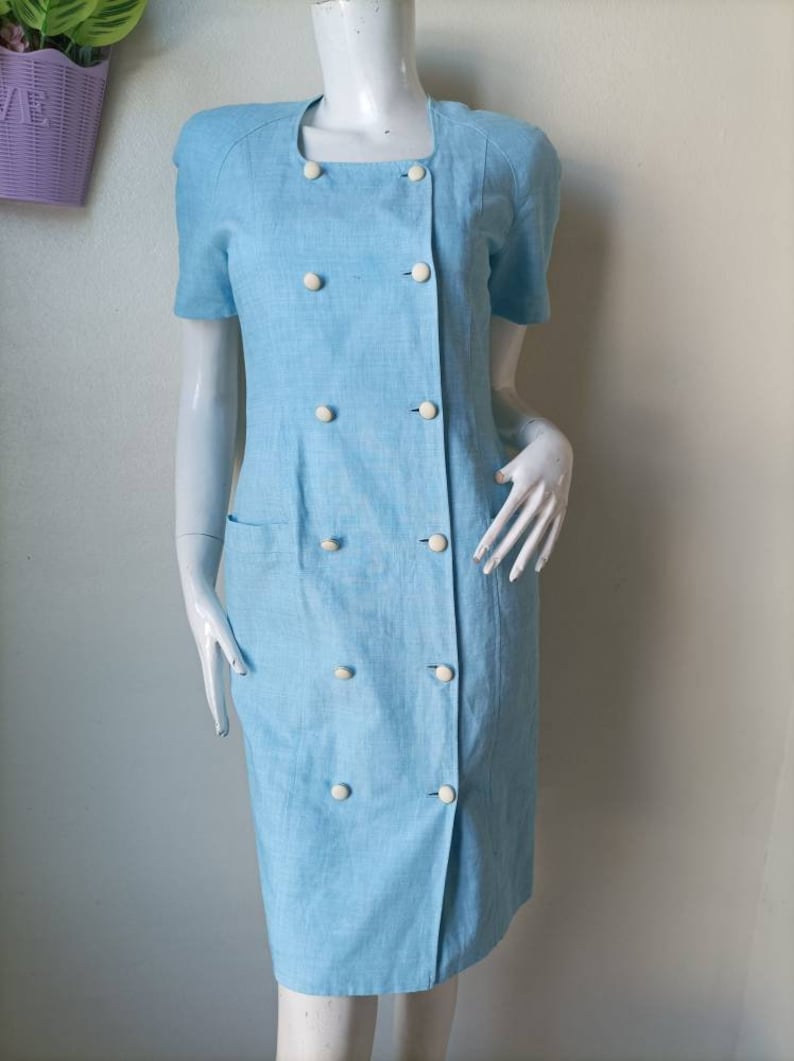 May include: A light blue dress with a double breasted button closure and short sleeves. The dress has a pocket on the left side and white buttons.