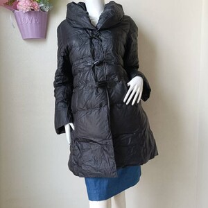 May include: Black puffer coat with a large collar and toggle fastenings. The coat is knee-length and has a quilted design. The mannequin is wearing a denim skirt. A purple basket with flowers is in the background.