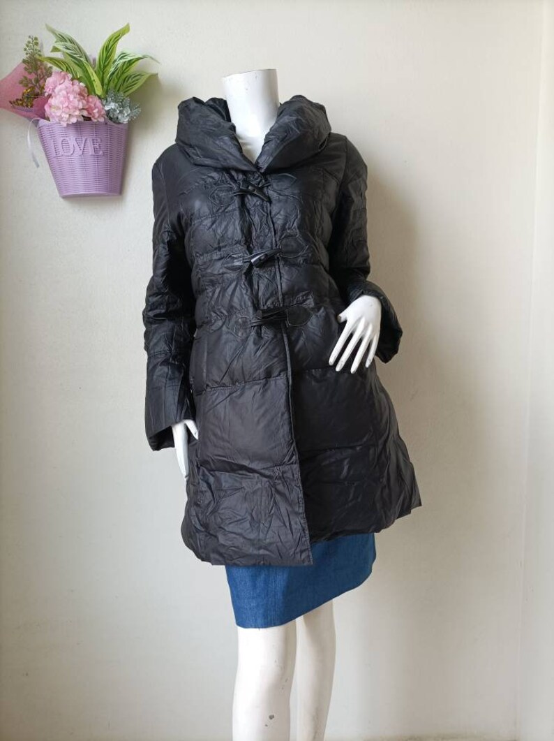 May include: Black puffer coat with a large collar and toggle fastenings. The coat is knee-length and has a quilted design. The mannequin is wearing a denim skirt. A purple basket with flowers is in the background.