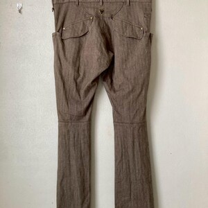 May include: Brown denim pants with a classic five-pocket design, featuring two front pockets and two back pockets. The pants are hanging on a wooden hanger against a white wall. The pants have a straight leg cut.