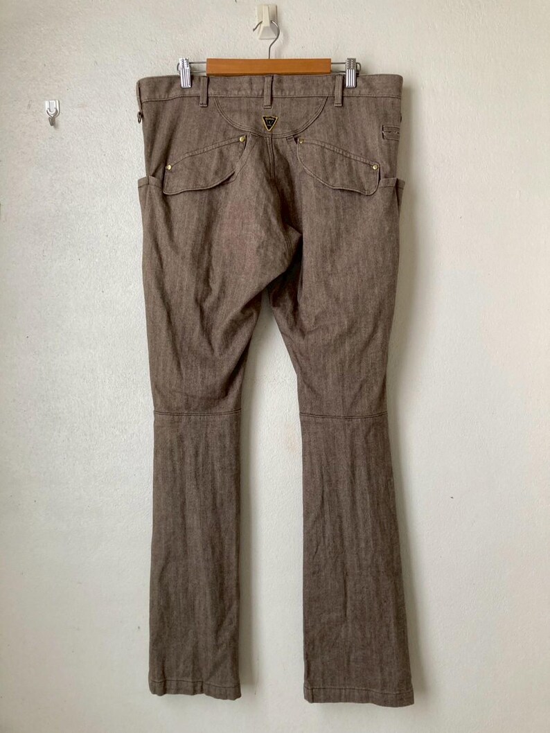 May include: Brown denim pants with a classic five-pocket design, featuring two front pockets and two back pockets. The pants are hanging on a wooden hanger against a white wall. The pants have a straight leg cut.