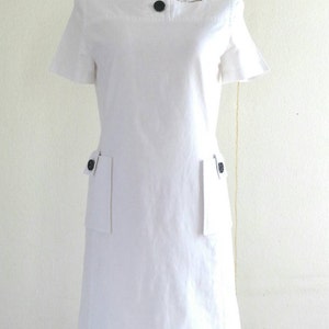 May include: A white short-sleeved dress with a square neckline and two patch pockets on the front. The dress has black buttons on the neckline and pockets.