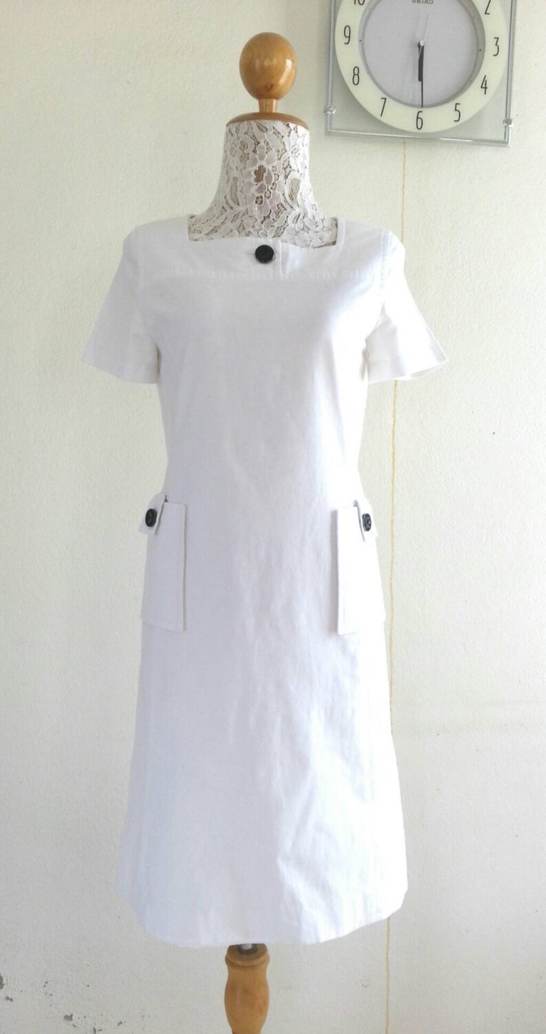 May include: A white short-sleeved dress with a square neckline and two patch pockets on the front. The dress has black buttons on the neckline and pockets.