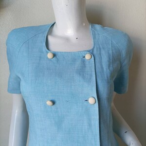 May include: A light blue linen dress with a double breasted closure and white buttons. The dress has short sleeves and a square neckline.