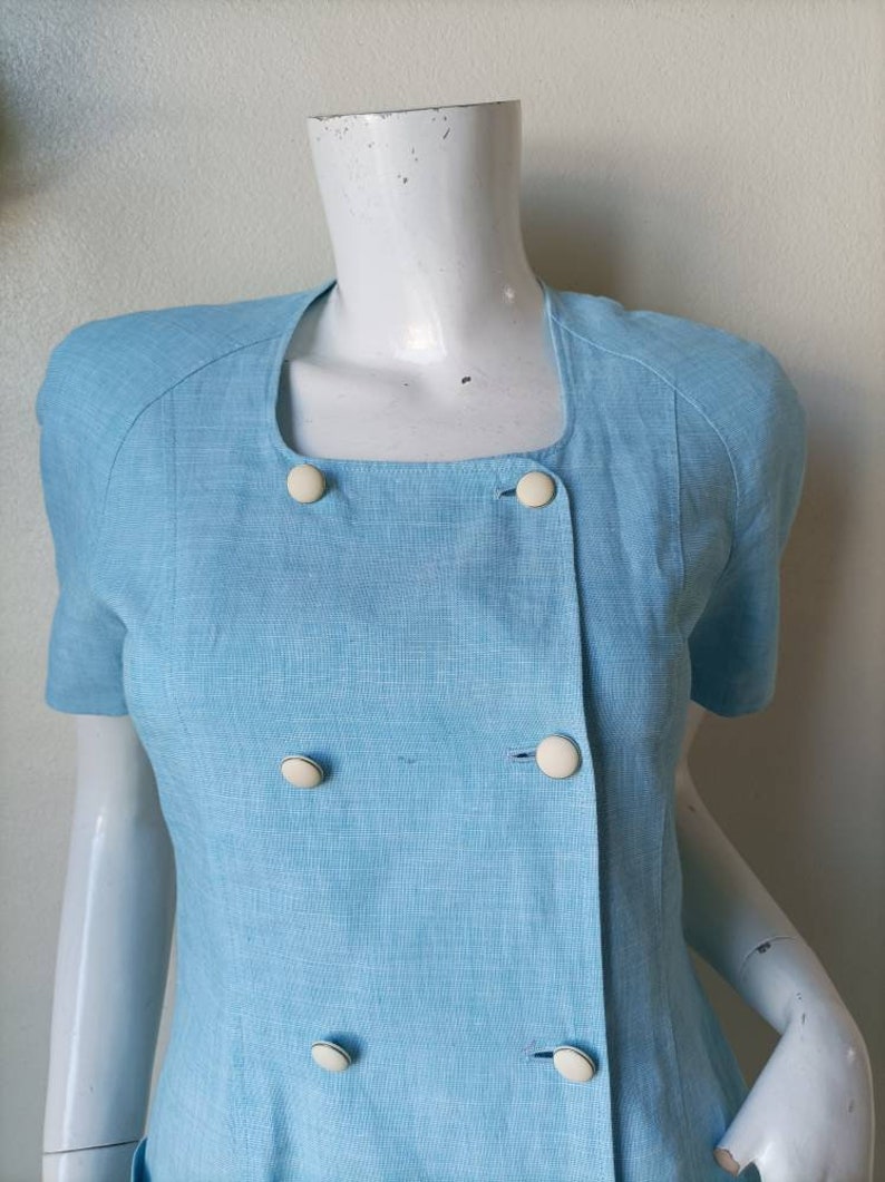 May include: A light blue linen dress with a double breasted closure and white buttons. The dress has short sleeves and a square neckline.