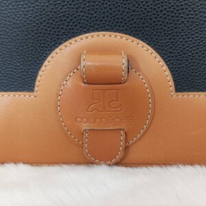 May include: A close-up of a brown leather purse with a black leather flap. The purse has a round, brown leather patch with the text "Courr&egrave;ges" and a logo in the center.