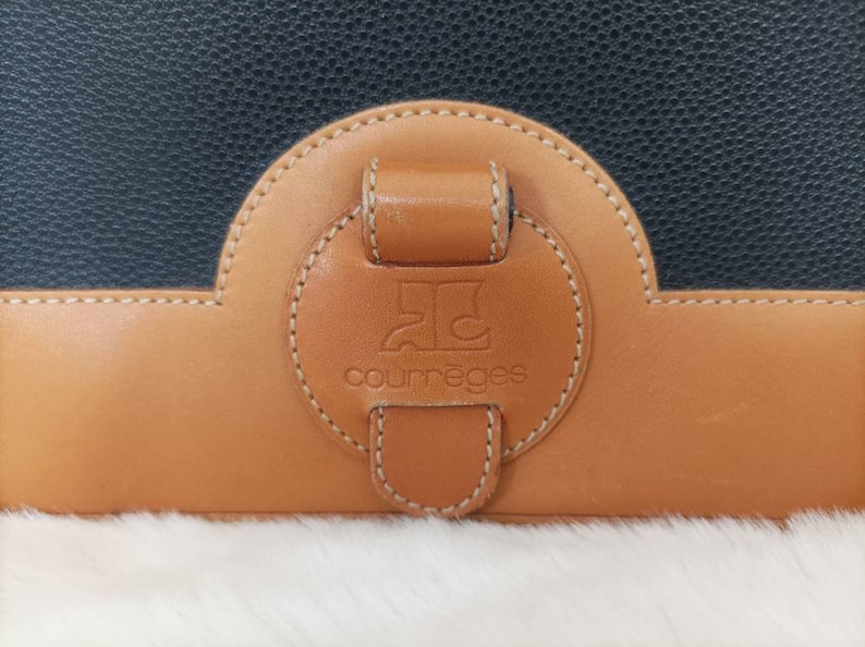 May include: A close-up of a brown leather purse with a black leather flap. The purse has a round, brown leather patch with the text "Courr&egrave;ges" and a logo in the center.