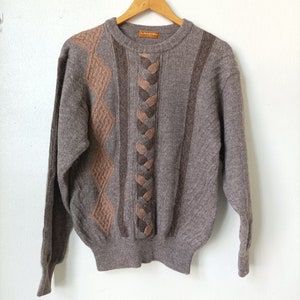 May include: A brown knitted sweater with a braided design and diamond pattern. The sweater has a crew neck and long sleeves. The brand name "LANCEL" is visible on the label.