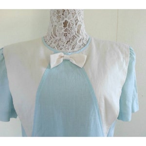 May include: A light blue dress with a white collar and a white bow. The dress has a unique design with white panels on the shoulders and a V-shaped neckline.