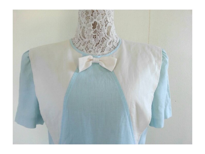 May include: A light blue dress with a white collar and a white bow. The dress has a unique design with white panels on the shoulders and a V-shaped neckline.