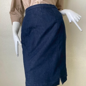 Puede incluir: Falda lápiz de mezclilla azul oscuro con abertura en el dobladillo. La falda se exhibe en un maniquí, combinada con una camisa abotonada color beige. La falda parece ser hasta la rodilla.