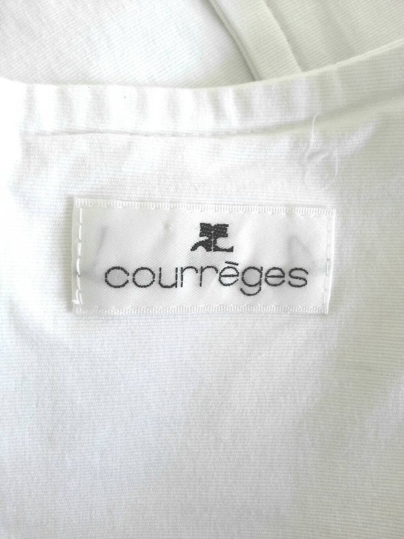May include: White clothing label with black text that reads "Courr&egrave;ges" with a stylized letter "C" above the word.