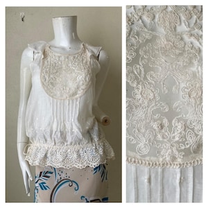 Op de afbeelding: Witte, mouwloze blouse met een delicate kanten overlay en een gerimpelde zoom. De kant heeft een bloemenpatroon.