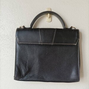 May include: A black leather briefcase with a top handle and a flap closure. The briefcase has a textured surface and is hanging on a hook.