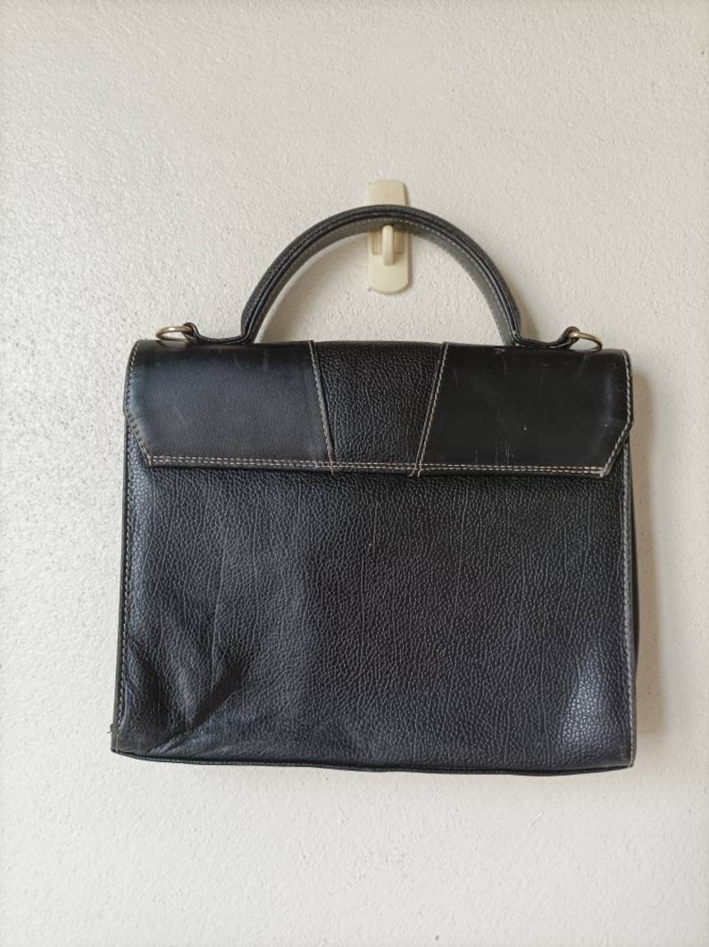 May include: A black leather briefcase with a top handle and a flap closure. The briefcase has a textured surface and is hanging on a hook.