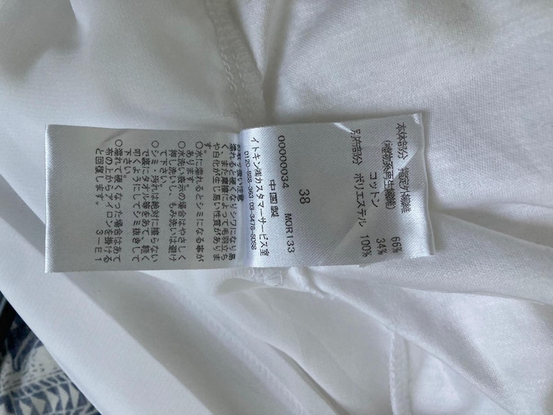 Peut inclure: Une &eacute;tiquette de tissu blanche avec du texte et des chiffres japonais, dont "38" et "MOR133". L'&eacute;tiquette indique la composition du tissu, avec des pourcentages de coton et de polyester. L'&eacute;tiquette est attach&eacute;e &agrave; un v&ecirc;tement blanc.