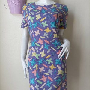 May include: A purple dress with a colorful abstract print of stars, lines, and crosses. The dress has short sleeves and a round neckline.