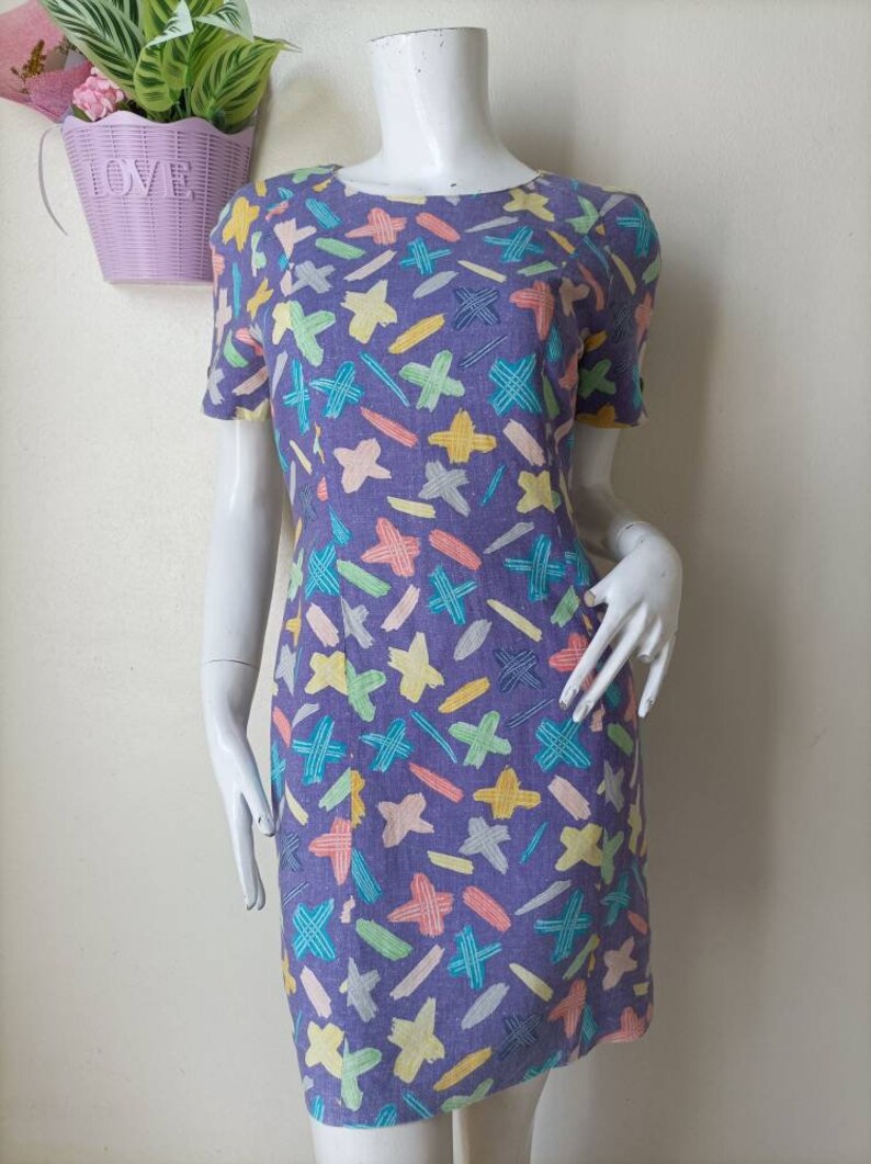 May include: A purple dress with a colorful abstract print of stars, lines, and crosses. The dress has short sleeves and a round neckline.
