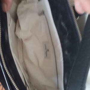 K&ouml;nnte beinhalten: Schwarze Ledertasche mit wei&szlig;em Stofffutter und Rei&szlig;verschluss. Das Futter hat ein kleines wei&szlig;es Etikett mit dem aufgedruckten Text "Coccinelle".
