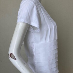 Peut inclure: Haut blanc &agrave; manches courtes avec des d&eacute;tails pliss&eacute;s. Le haut pr&eacute;sente une taille ajust&eacute;e et un design superpos&eacute; sur le corsage. Les manches sont courtes et le tissu semble l&eacute;ger. Le haut est pr&eacute;sent&eacute; sur un mannequin.