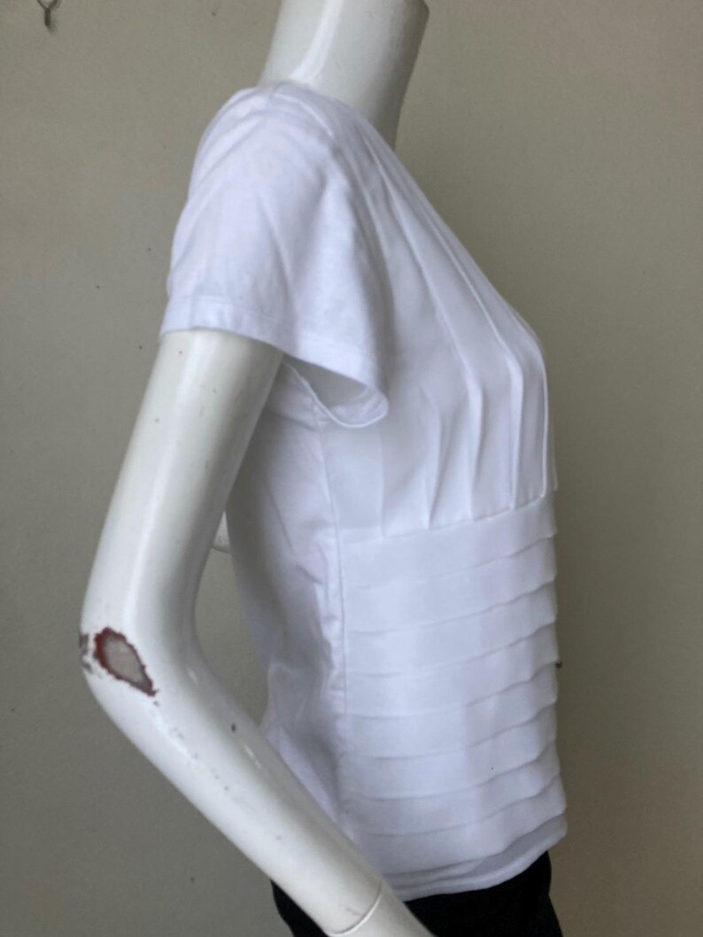 Peut inclure: Haut blanc &agrave; manches courtes avec des d&eacute;tails pliss&eacute;s. Le haut pr&eacute;sente une taille ajust&eacute;e et un design superpos&eacute; sur le corsage. Les manches sont courtes et le tissu semble l&eacute;ger. Le haut est pr&eacute;sent&eacute; sur un mannequin.