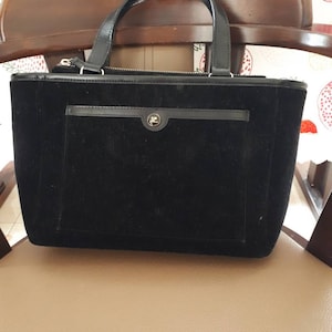 K&ouml;nnte beinhalten: Eine schwarze Cordtasche mit Rei&szlig;verschluss oben und einer kleinen Tasche auf der Vorderseite. Die Tasche hat ein goldenes Logo auf der vorderen Tasche.