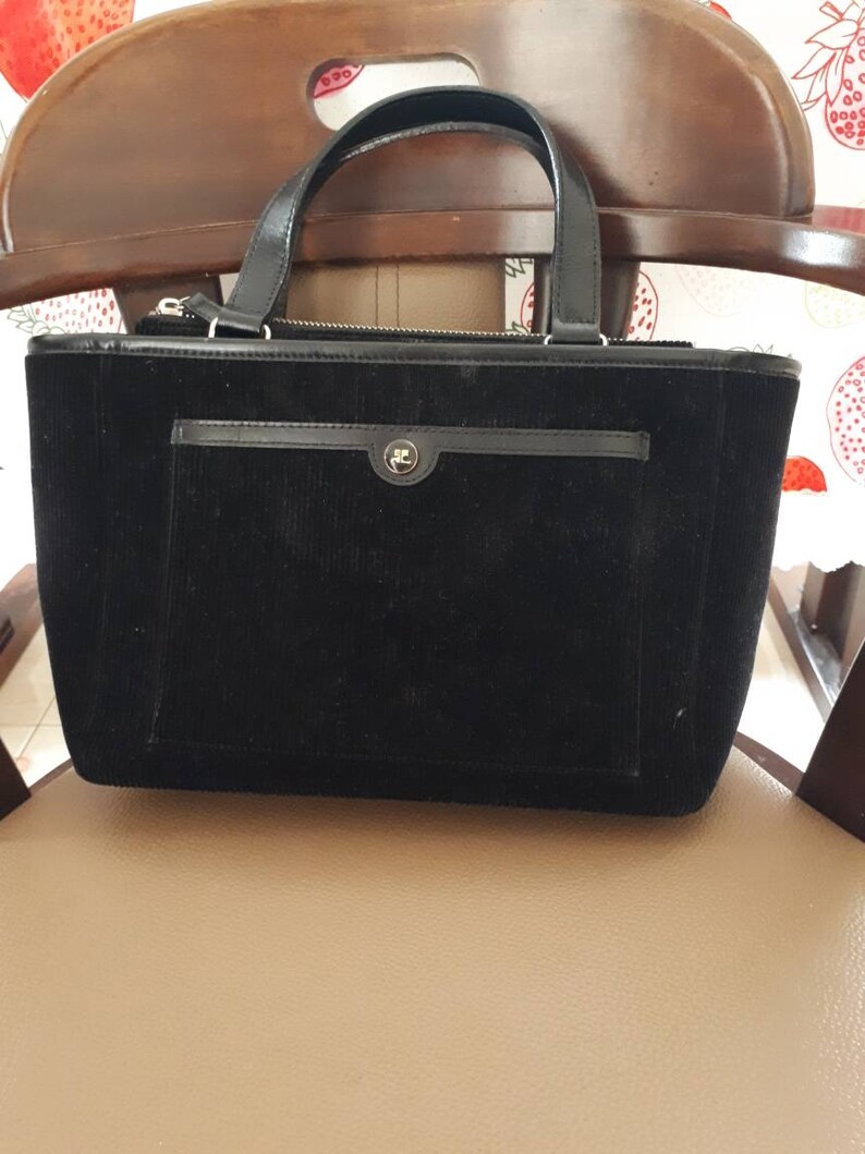 K&ouml;nnte beinhalten: Eine schwarze Cordtasche mit Rei&szlig;verschluss oben und einer kleinen Tasche auf der Vorderseite. Die Tasche hat ein goldenes Logo auf der vorderen Tasche.