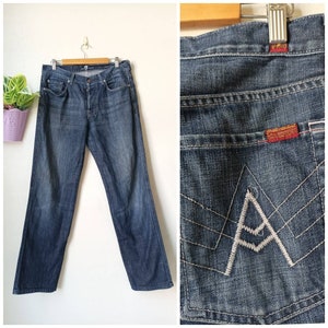 Puede incluir: Un par de pantalones vaqueros de color azul oscuro con un detalle de costura azul claro en el bolsillo trasero. El bolsillo trasero tiene una letra 'A' bordada en blanco.