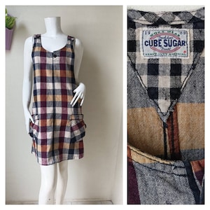 May include: A black, brown, and white plaid overall dress with two pockets on the front. The dress has a label that reads "Work Wear 1989 Tradition Cube Sugar Trust Heavy Duty Material" with a red star.