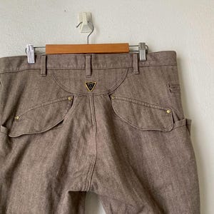 May include: Brown denim pants with a unique pocket design and gold-colored rivets. The pants feature a distinctive back yoke and a small gold emblem. They are hanging on a wooden hanger against a white wall.