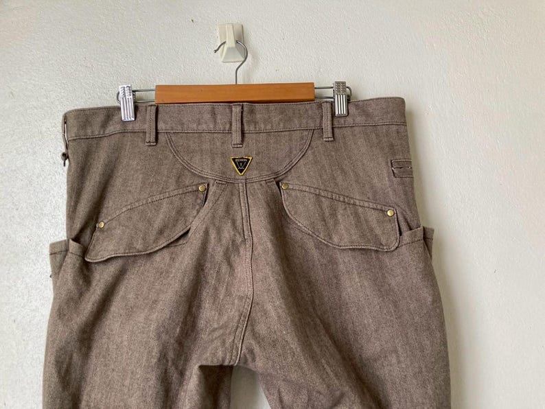 May include: Brown denim pants with a unique pocket design and gold-colored rivets. The pants feature a distinctive back yoke and a small gold emblem. They are hanging on a wooden hanger against a white wall.