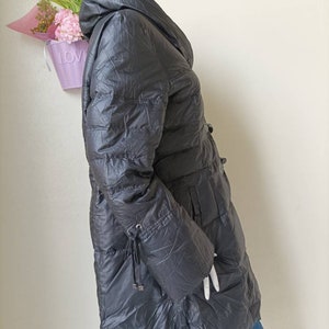 May include: A dark grey puffer coat with a hood, displayed on a mannequin. The coat features a quilted design, a cinched waist, and drawstring details on the sleeves. The coat is long, extending to the mid-thigh.